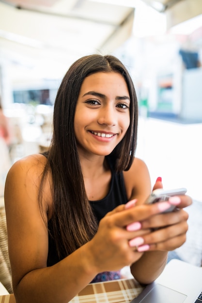 Young woman in a cafe reading a text message from her phone. Latin female sitting at cafe table with laptop and using smart phone.