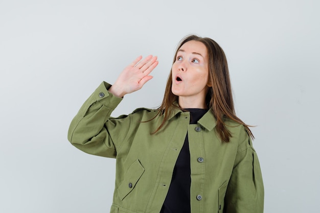 Young woman calling someone with loud voice in green jacket and looking focused. front view.