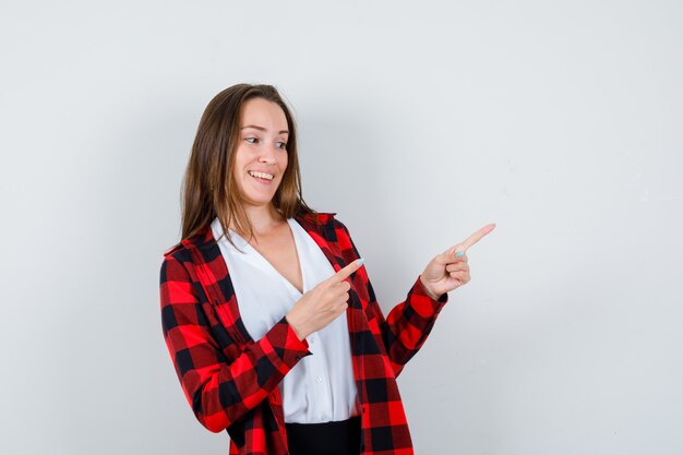 Young woman in casual clothes pointing at upper right corner and looking jovial , front view.