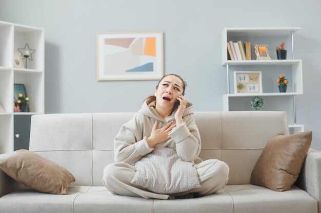 Молодая женщина в повседневной одежде сидит на диване в домашнем интерьере и выглядит взволнованной и нервной, разговаривая по мобильному телефону и собираясь плакать
