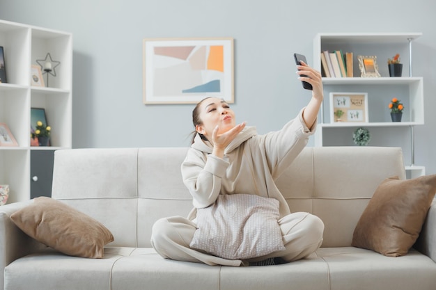 Молодая женщина в повседневной одежде сидит на диване в домашнем интерьере с помощью смартфона и делает видеозвонок, отправляя воздушный поцелуй счастливым и позитивным