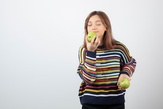 Young woman in casual outfit eating green apple on white background. 