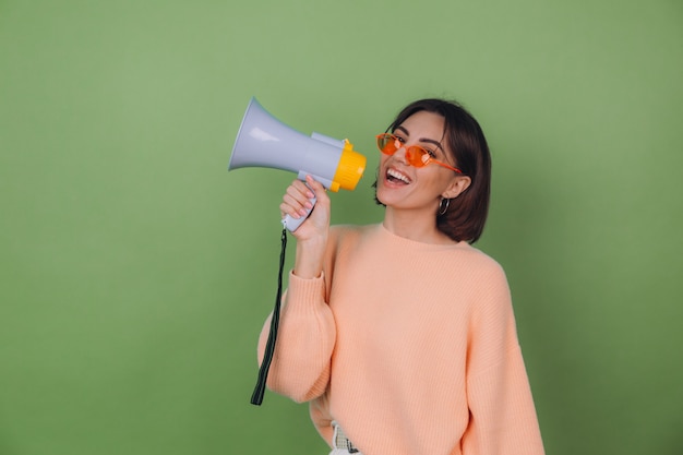 Young woman in casual peach and orange eyeglasses sweater isolated on green olive wall happy screaming in megaphone copy space
