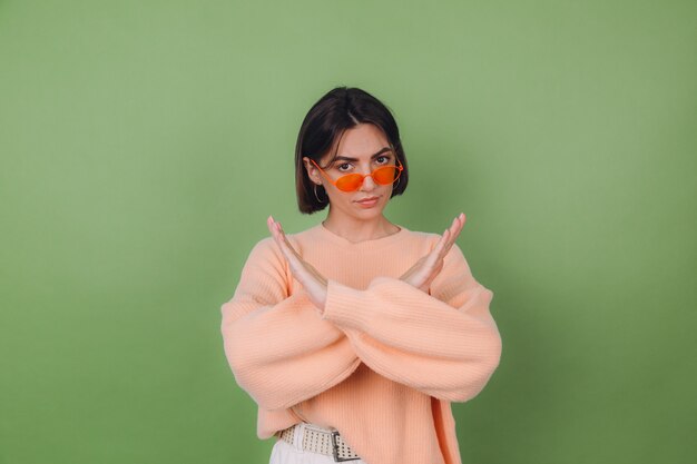 Young woman in casual peach and orange eyeglasses sweater isolated on green olive wall serious showing stop gesture with crossed hands copy space