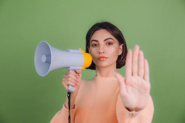 Young woman in casual peach sweater isolated on green olive color wall. unhappy serious with megaphone doing stop sing with palm of the hand