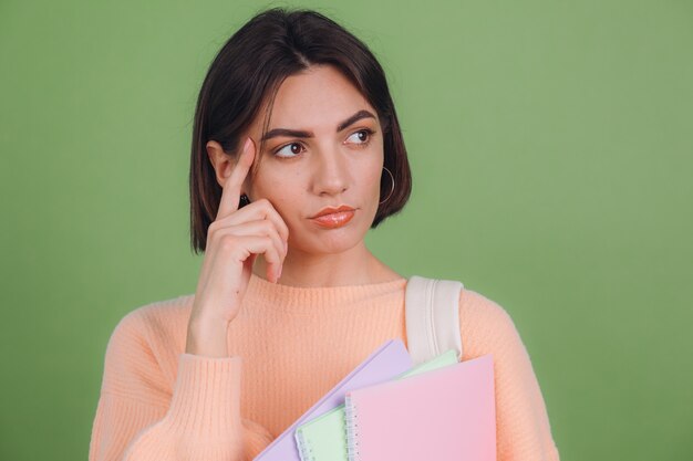 緑のオリーブ色の壁に分離されたカジュアルな桃のセーターの若い女性