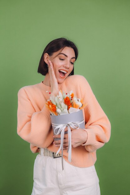 녹색 올리브 벽에 고립 된 캐주얼 복숭아 스웨터에 젊은 여자 목화 꽃 라든지 밀과 lagurus의 오렌지 흰색 꽃 상자 구성 선물 행복 놀란 놀란