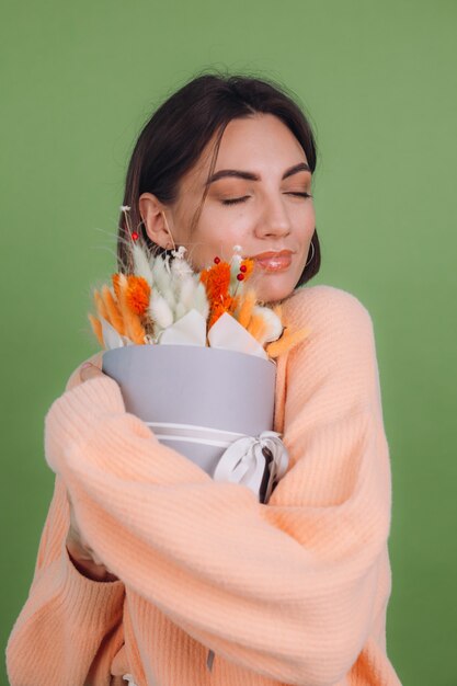 緑のオリーブの壁に分離されたカジュアルな桃のセーターの若い女性は、ギフトのための綿の花カスミソウ小麦とラグラスのオレンジ色の白いフラワーボックスの構成を保持します幸せな驚き驚き