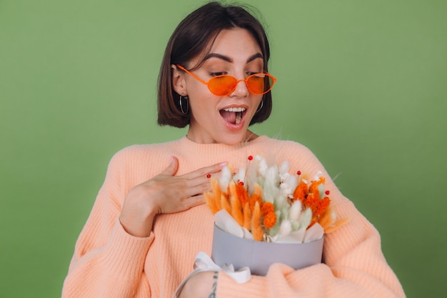 녹색 올리브 벽에 고립 된 캐주얼 복숭아 스웨터에 젊은 여자는 목화 꽃, 라든지, 밀, lagurus의 오렌지 흰색 꽃 상자 구성을 잡고 행복 놀라게 선물