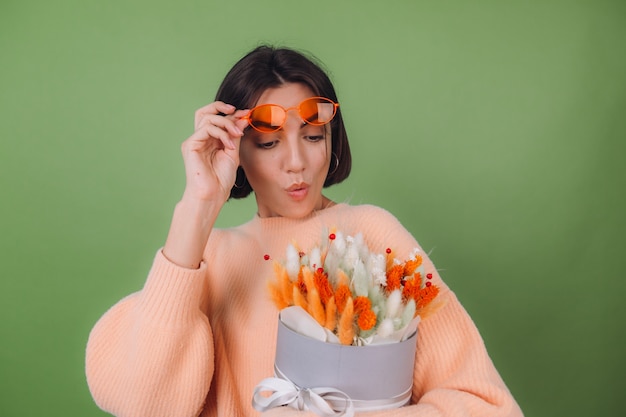 Young woman in casual peach sweater  isolated on green olive wall  hold  orange white flower box composition of cotton flowers, gypsophila, wheat and lagurus for a gift happy surprised