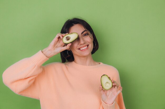 Young woman in casual peach sweater isolated on green olive wall holding  avocado, health and skin care concept, copy space