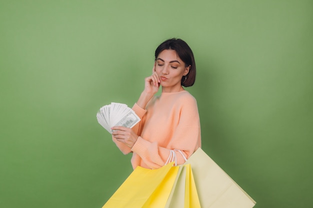 Молодая женщина в повседневном персиковом свитере изолирована на зеленой оливковой стене, держа веер с деньгами и сумками для покупок в 100 долларов, думая позитивно, улыбаясь, копируя пространство