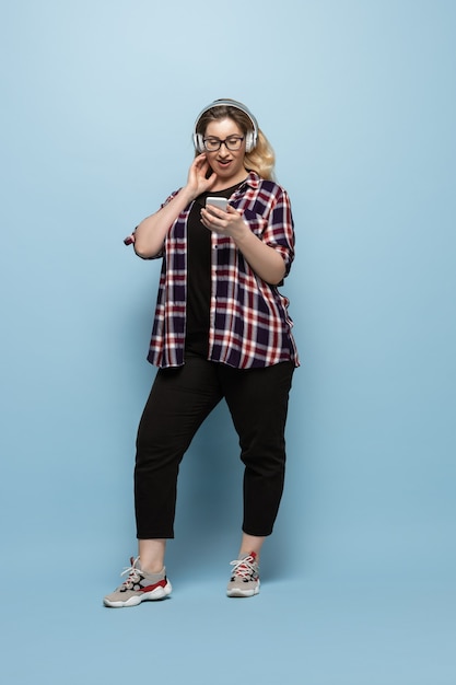 Free photo young woman in casual wear with smartphone and headphones on blue wall. body positive character, feminism, loving herself, beauty concept. plus size beautiful businesswoman. inclusion, diversity.