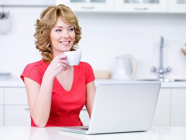 Young woman in casuals with cup of coffee and laptop in the kitchen