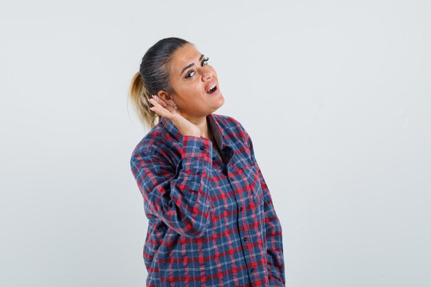 Young woman in checked shirt holding hand near ear as trying to listen something and looking focused , front view.