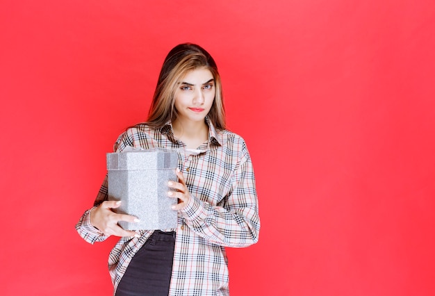 Young woman in checked shirt holding a silver gift box and looks confused and thoughtful