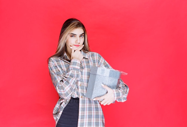 Young woman in checked shirt holding a silver gift box and looks confused and thoughtful
