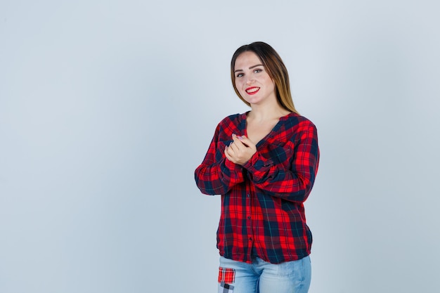 Young woman in checked shirt, jeans clasping hands, smiling and looking cheery , front view.