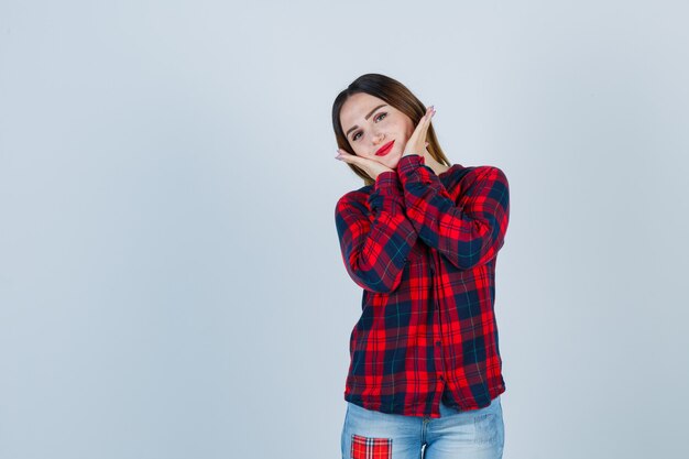 Young woman in checked shirt, jeans pillowing face on hands and looking cute , front view.