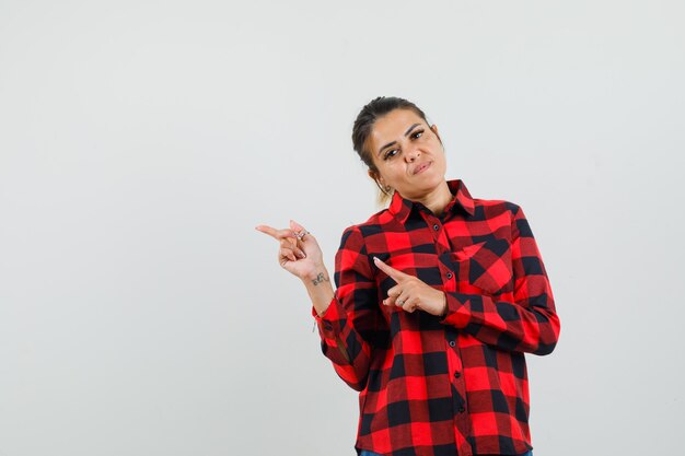 Young woman in checked shirt pointing at left corner and looking confident , front view.