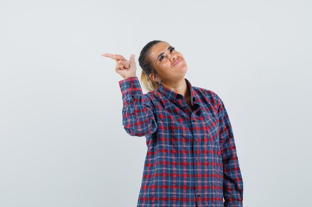 Free photo young woman in checked shirt pointing left with index finger and looking pretty , front view.