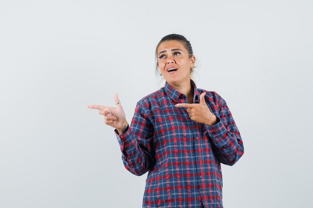 Young woman in checked shirt pointing left with index fingers and looking pretty , front view.