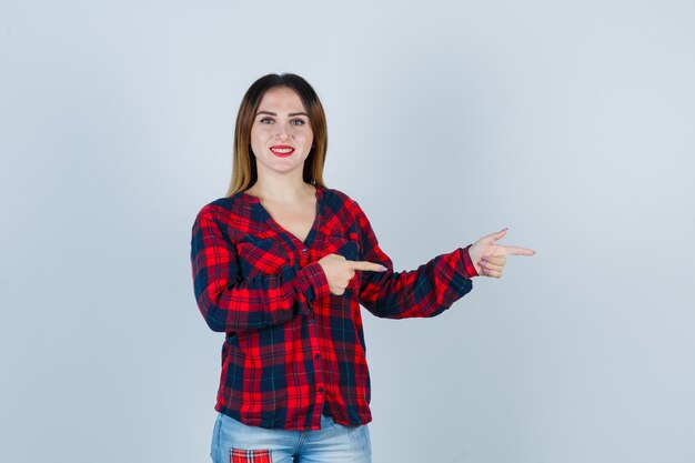 Young woman in checked shirt pointing right and looking cheerful , front view.