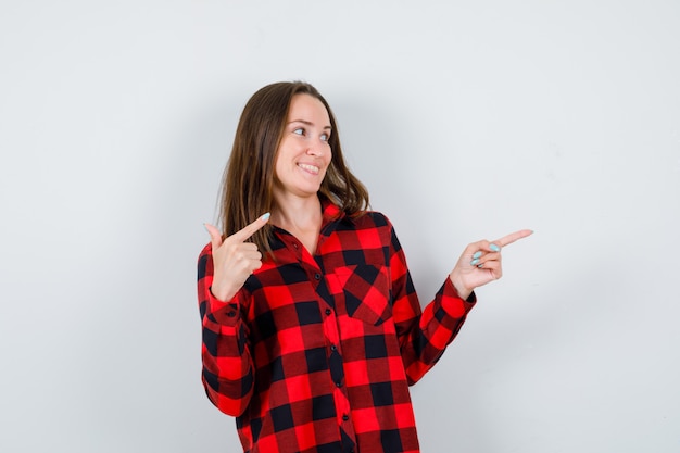 Young woman in checked shirt pointing right with index fingers and looking cheery , front view.