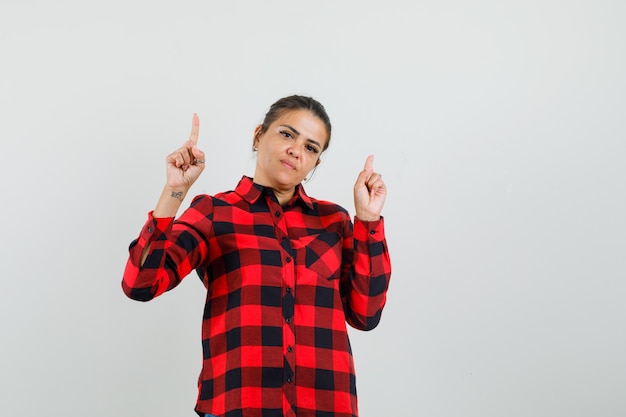 Young woman in checked shirt pointing up and looking confident , front view.