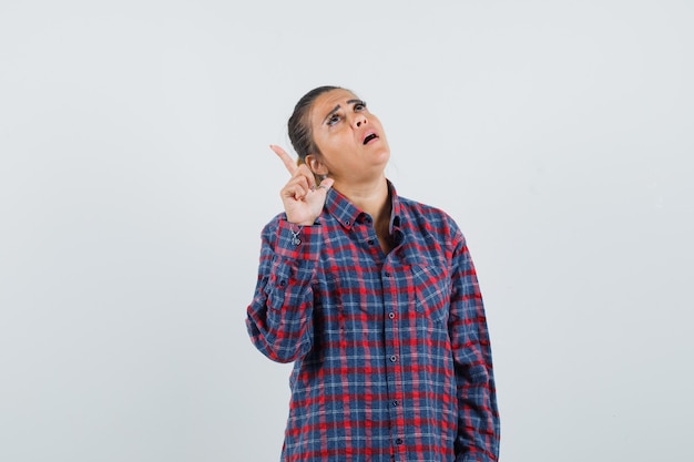 Young woman in checked shirt pointing up with index finger and looking pretty , front view.