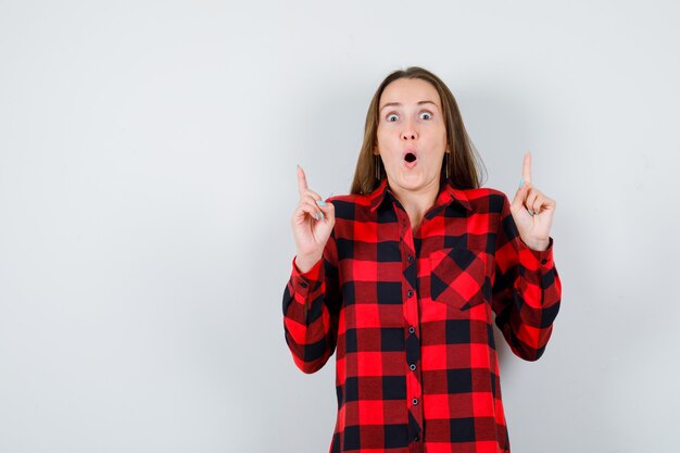 Young woman in checked shirt pointing up with index fingers and looking shocked , front view.