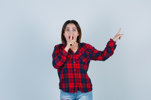 Young woman in checked shirt showing silence gesture, pointing up and looking excited , front view.