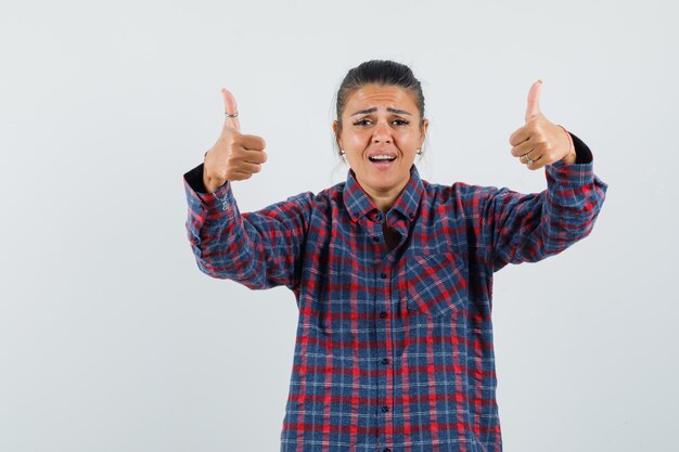 Young woman in checked shirt showing thumbs up and looking happy , front view.