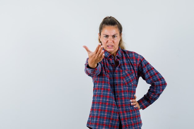 Giovane donna in camicia a quadri che allunga la mano come chiamare qualcuno mentre tiene un'altra mano sulla vita e sembra arrabbiata