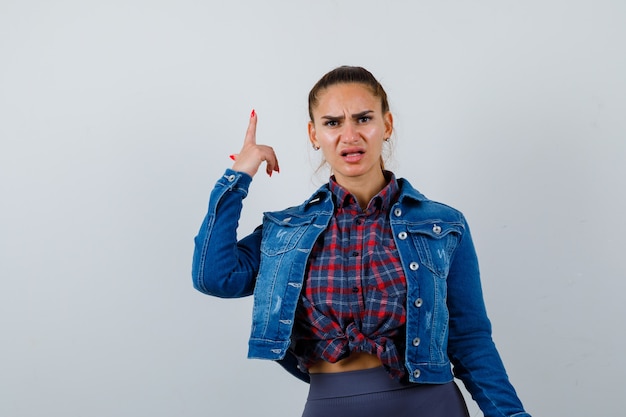 Free photo young woman in checkered shirt, jean jacket pointing up and looking resentful , front view.