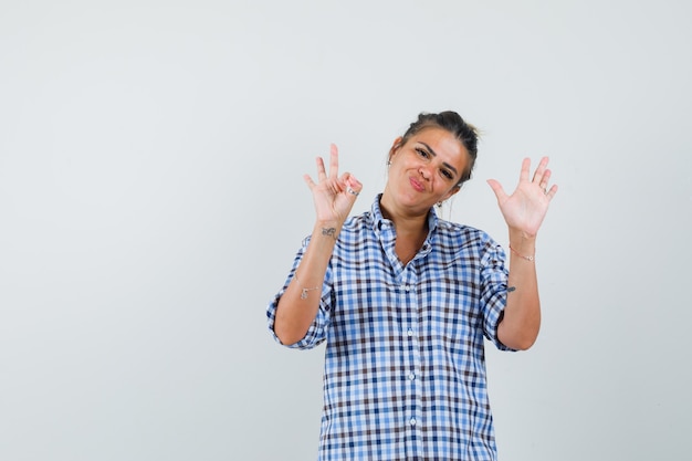 Young woman in checkered shirt showing ok gesture while raising hand up and looking hopeful.