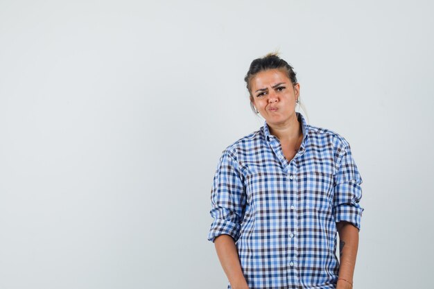 Young woman in checkered shirt standing while pouting lips and looking unhappy.