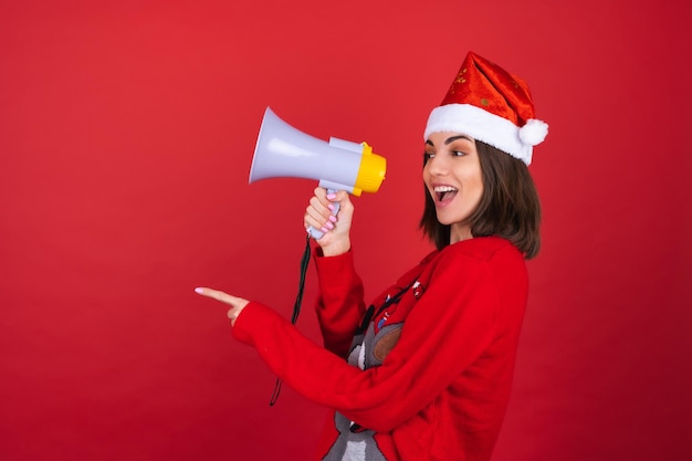 Foto gratuita la giovane donna con un maglione natalizio e un cappello da babbo natale con un megafono è eccitata, allegra, punta il dito a sinistra