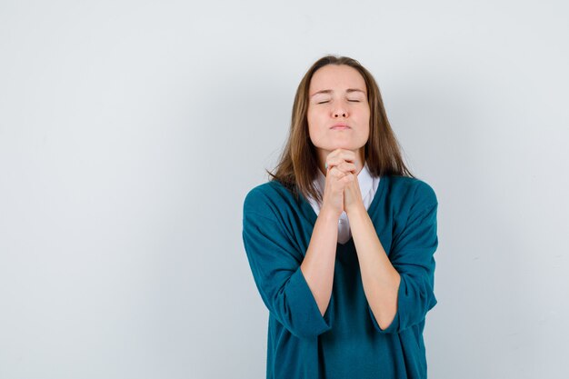 Young woman clasping hands in pleading gesture in sweater over white shirt and looking hopeful. front view.