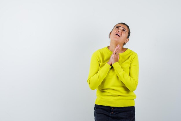 Young woman clasping hands in prayer position in yellow sweater and black pants and looking dismal 