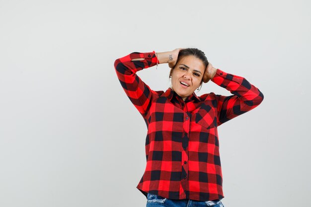 Young woman clasping head with hands in checked shirt, shorts and looking cute , front view.