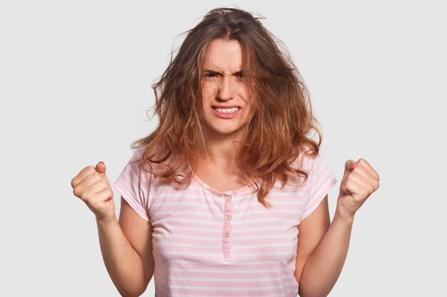Young woman clenches fists with anger, looks irritated, has mess on head, feels furious when hears sound of alarm clock