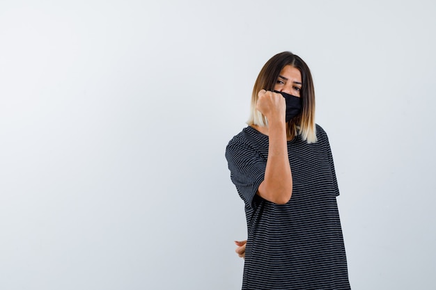 Giovane donna che stringe il pugno in abito nero, maschera nera e sembra potente. vista frontale.