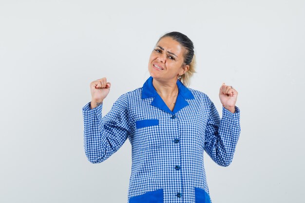 Young woman clenching fists in blue gingham pajama shirt and looking pretty , front view.