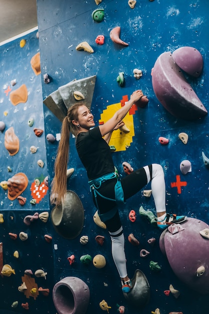 Молодая женщина поднимается на высокую искусственную стену для скалолазания в помещении