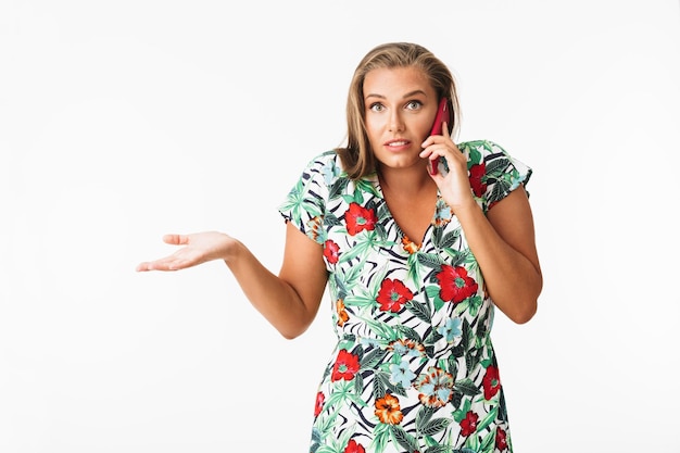 Young woman in colorful dress amazedly looking in camera while talking on cellphone over white background