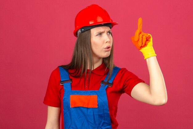 건설 유니폼 장갑과 빨간색 안전 헬멧에 젊은 여자는 새로운 아이디어가 서 있고 어두운 분홍색 배경에 손가락으로 가리키는