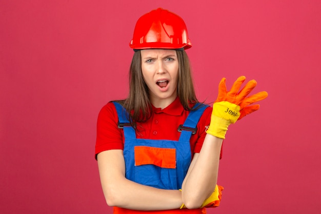 Молодая женщина в перчатках строительной формы и красном защитном шлеме, заставляющая задавать вопрос о чувстве недоумения и негодования на темно-розовом фоне