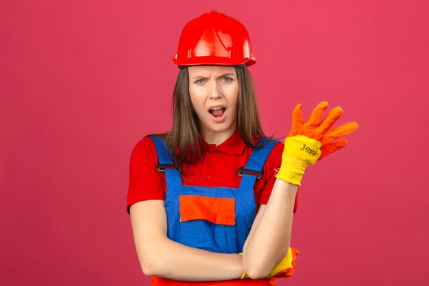 建設の均一な手袋と質問のジェスチャーを作る赤い安全ヘルメットの若い女性は暗いピンクの背景に当惑して憤慨している感じ