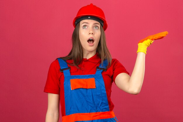 暗いピンクの背景に上げられた手で立っている驚きの表情でショックを受けた建設制服手袋と赤い安全ヘルメットの若い女性